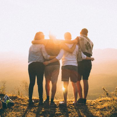 Four friends stand with their arms around each other as they watch the sunset. Their backs are facing the camera. It's okay to ask for help.