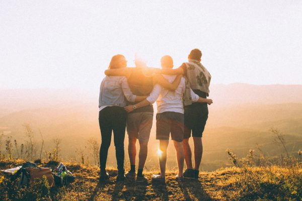 Four friends stand with their arms around each other as they watch the sunset. Their backs are facing the camera. It's okay to ask for help.