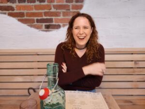 Christy sitting at a table, laughing. An exposed brick wall is visible in the background, partially plastered over. Christy appears to be at a restaurant; a water bottle and a menu are on the table.