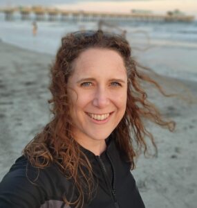 Christy Nicholson on a beach, wearing black. She is facing the camera, and a pier is in the background.